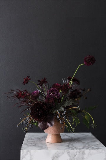 Pink Tarte footed pot holding a dark burgundy floral arrangement on a marble pedestal