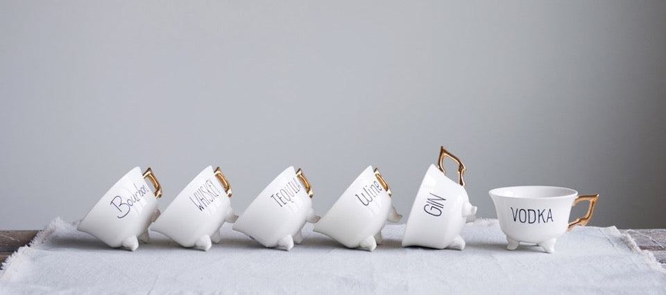 Six tea cups with gold handles tilted on table.  Each tea cup has black text with Wine, Vodka, Gin, Tequila, Whiskey, or Bourbon on side.