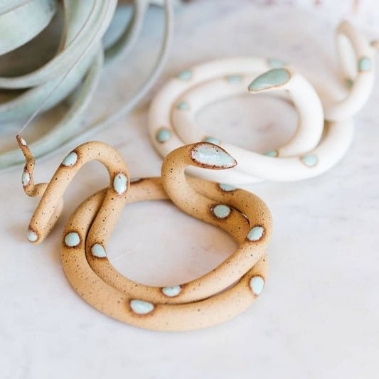 Two coiled ceramic snakes, one speckled tan and one white, both with mint green spots, on white marble